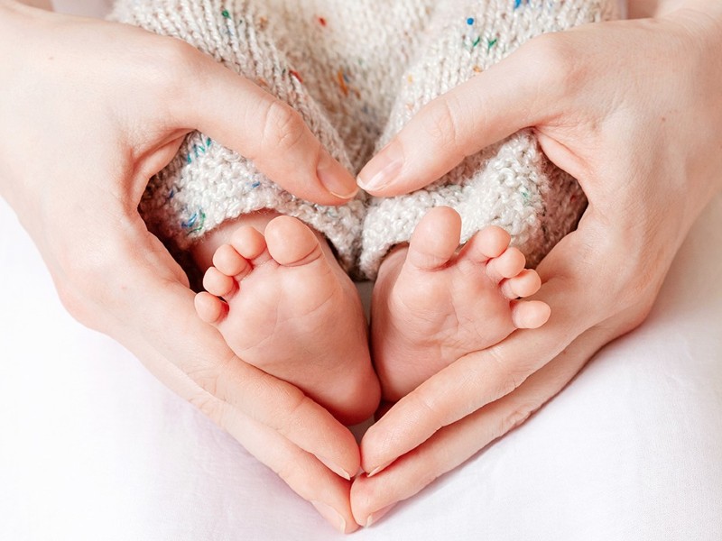Foto de duas mãos femininas formando um coração em volta dos pezinhos de um bebê, que veste uma calça de tricô clara